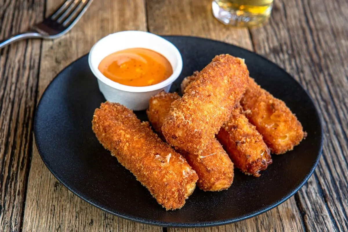 A black plate topped with fried cheese curds next to a cup of dipping sauce.