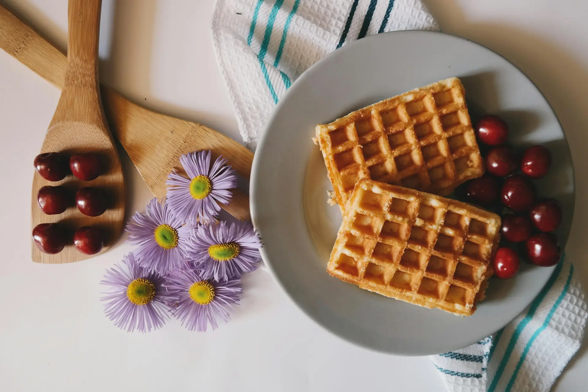 On a plate there are two pieces of waffles along with some red cherries. Next to the plate there is some spoons, and flowers.