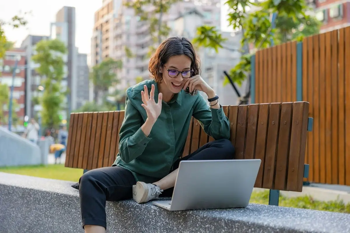 A person waving at a laptop, possibly saying hello to somebody, while sitting in a park.
