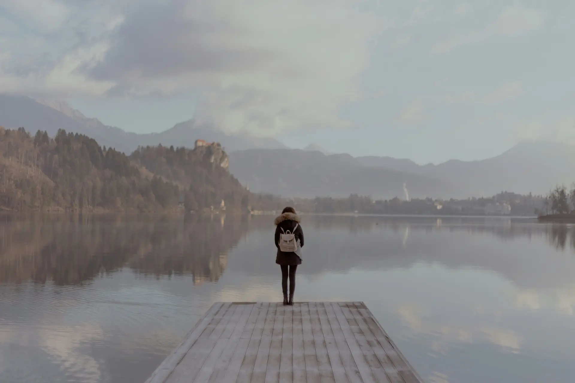 Woman standing on deck of a lake. Mountains and forests can be seen in the distance.