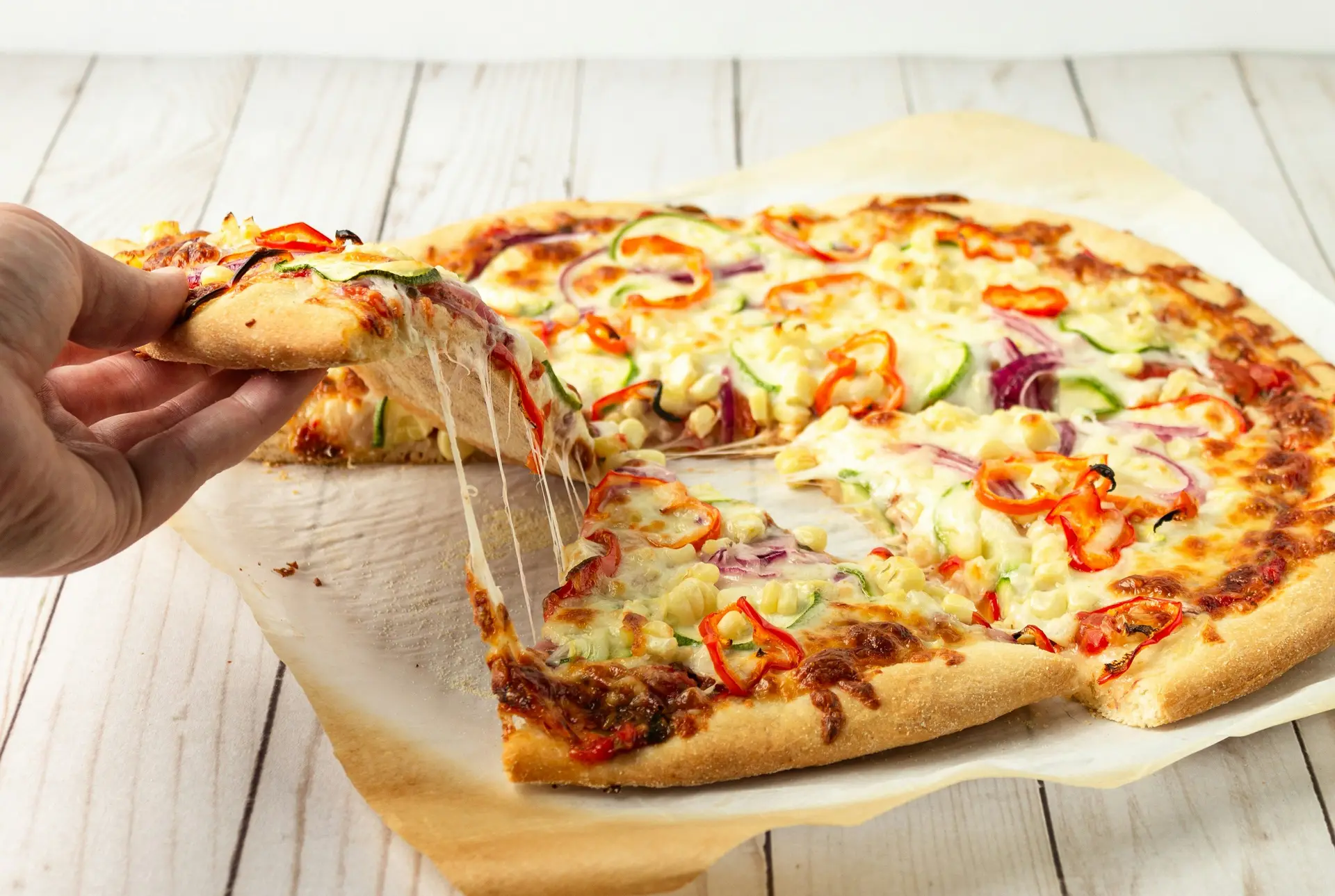 A person taking a slice of cheese pizza. The pizza is placed on a wooden table.