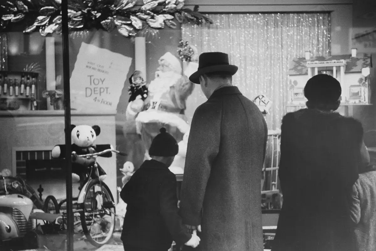 A family with a child standing before a toy shop with bicycle, teddy bears, and other toys on display.. It's a vintage black and white photo.
