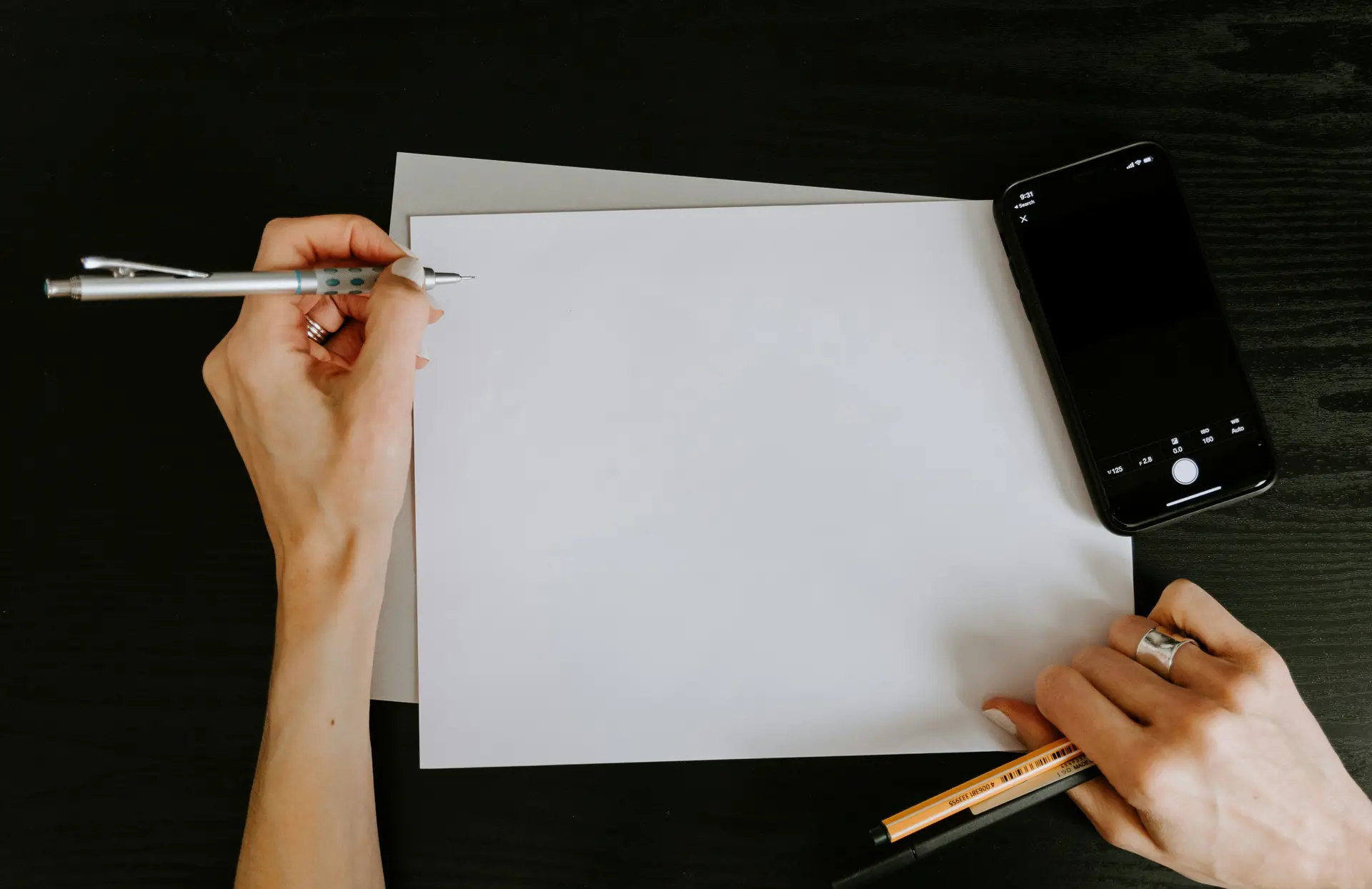 A person holding a pen over a empty white page. There's a mobile next to the pad.