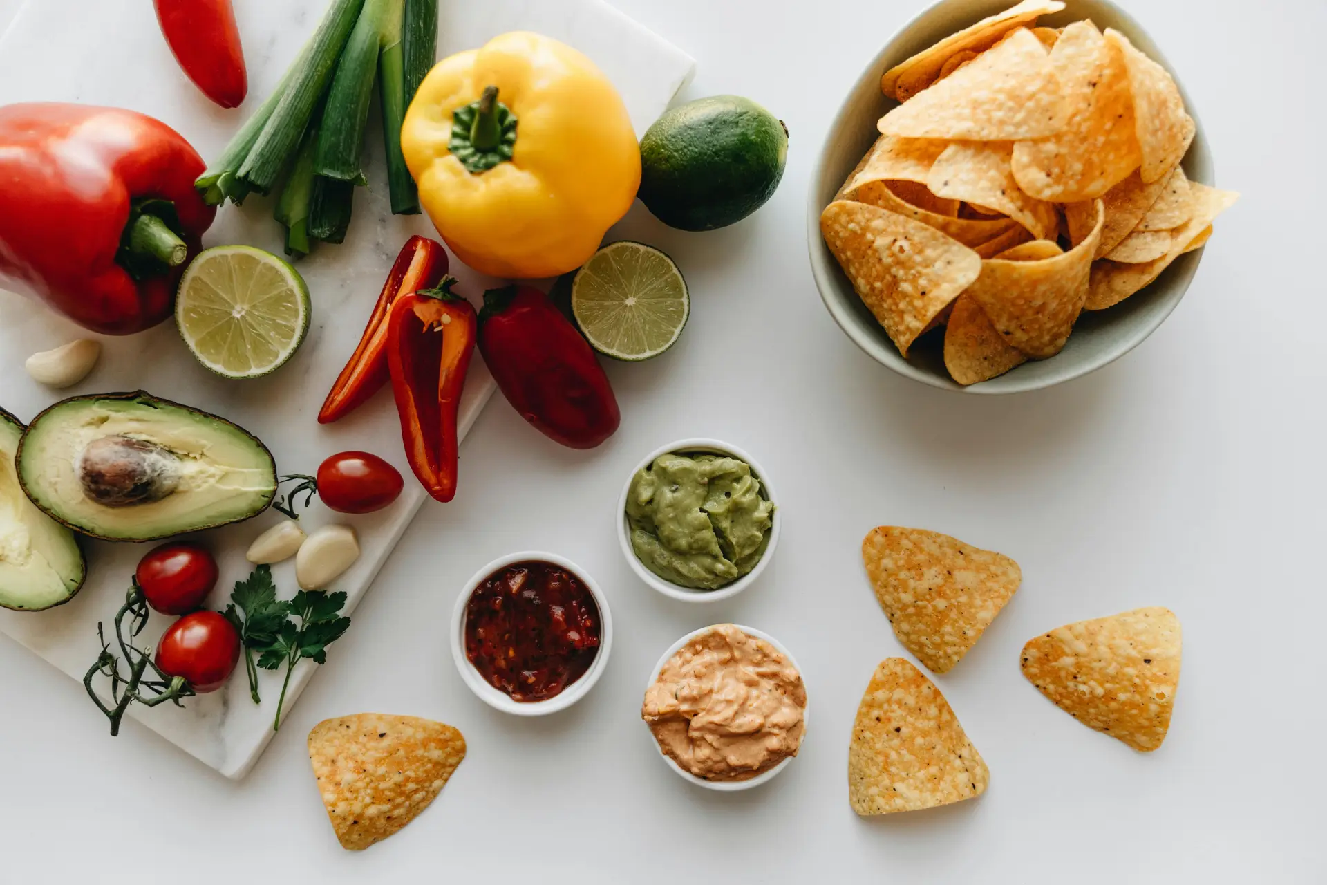 Potato chips, three types of dips, yellow capsicum, avocado, cherry tomatoes, red chilies, lemon and other ingredients are decorated on a white platform.