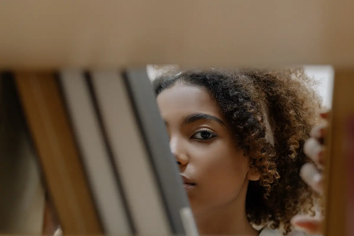 Close-up portrait of a woman peeking through books on a library shelf.
