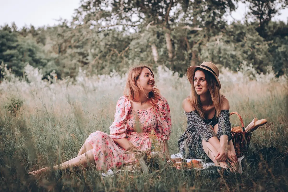 Two women enjoy a summer picnic in a scenic grass field, dressed in floral dresses, embodying friendship and style.