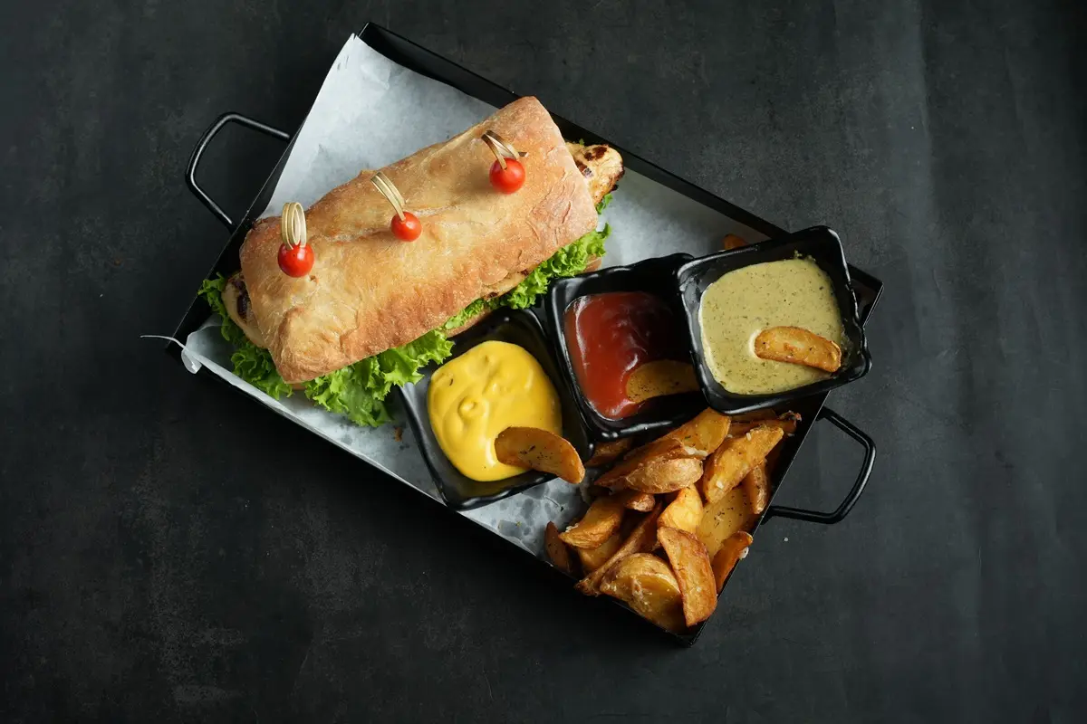 A gourmet sandwich served with fries and sauces on a black tray.
