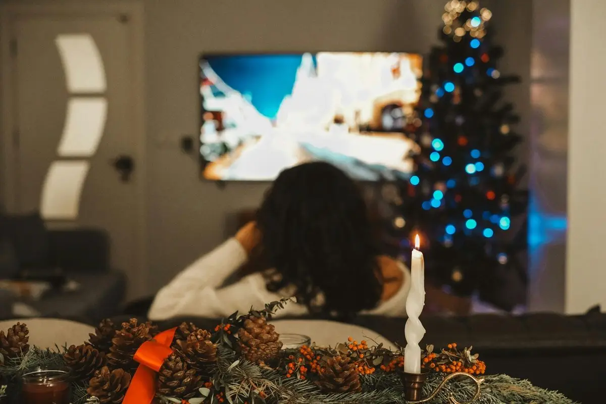 A person watching tv while sitting on a sofa in the living room that has a Christmas tree.