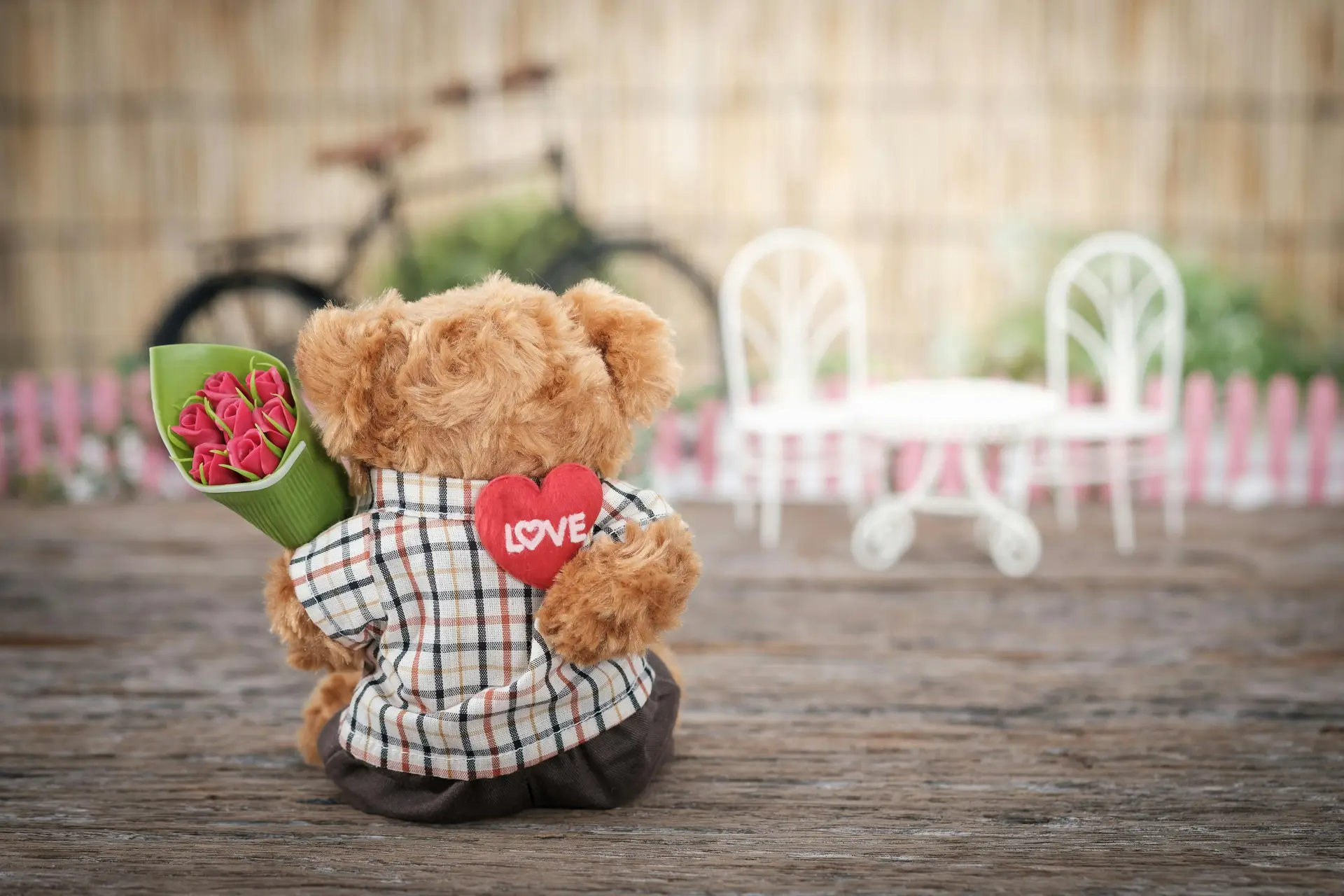 Brown bear plush toy holding red Rose flower while stting on a wooden surface.