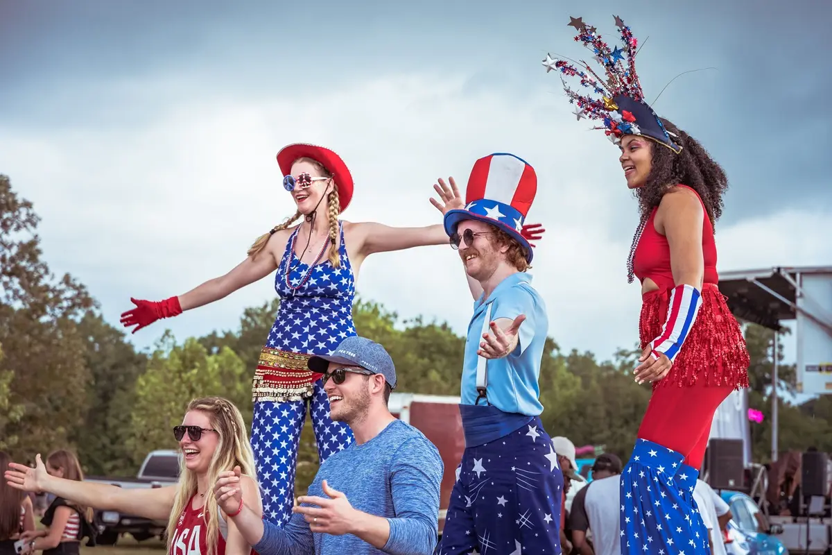 Colorful outdoor event featuring performers on stilts in patriotic attire, smiling and engaging with a cheerful crowd.