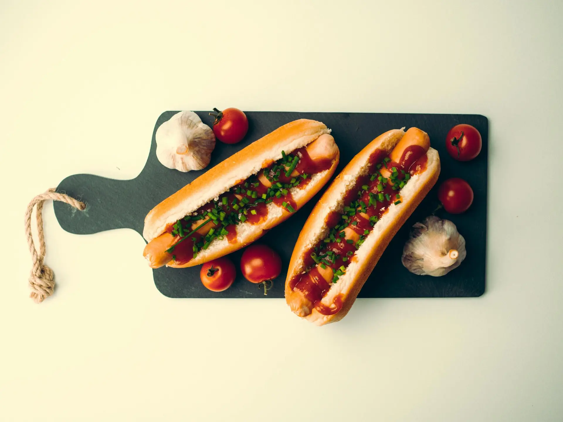 Two hot dogs on a chopping board along with garlic and tomateos.