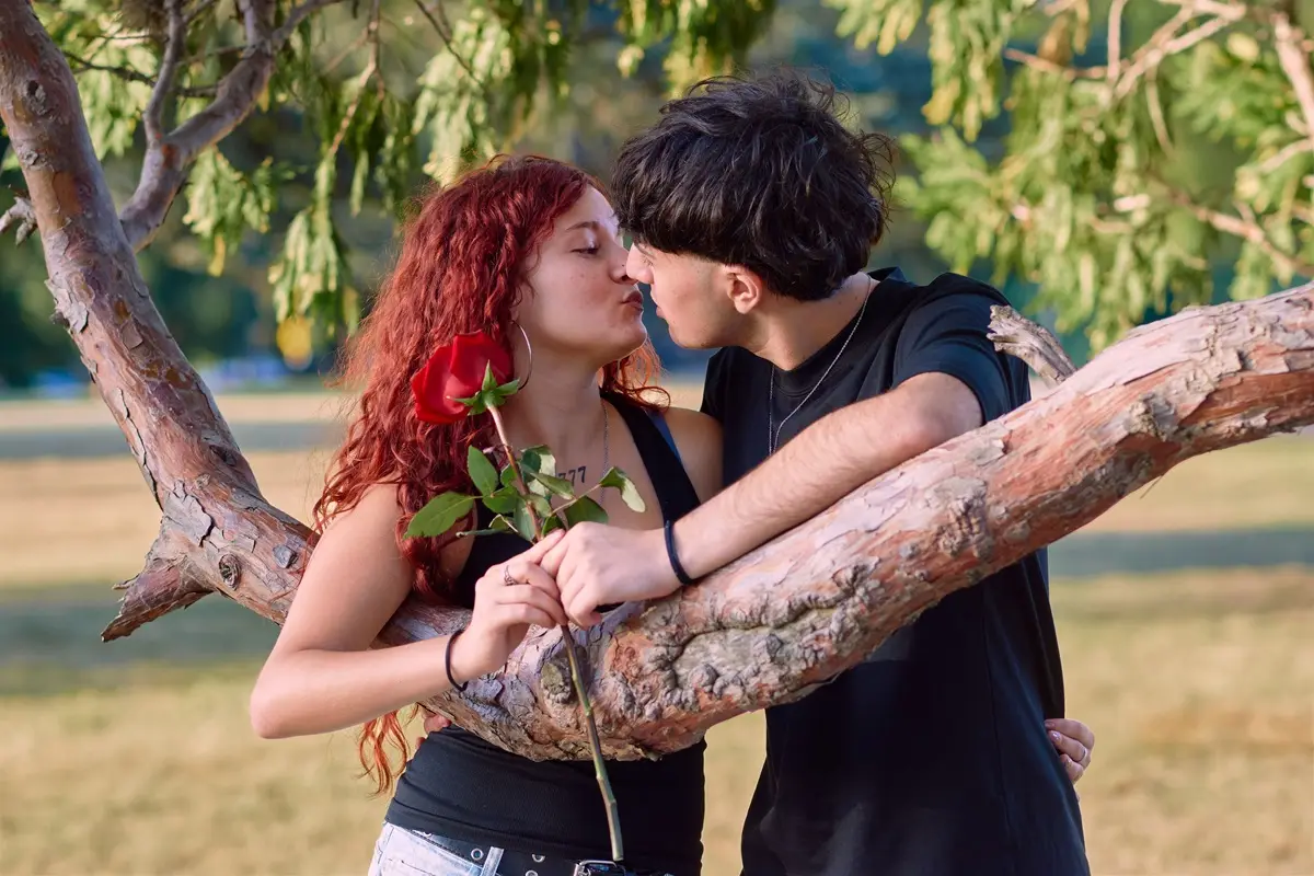 Young couple sharing a romantic kiss beneath a tree, holding a red rose.