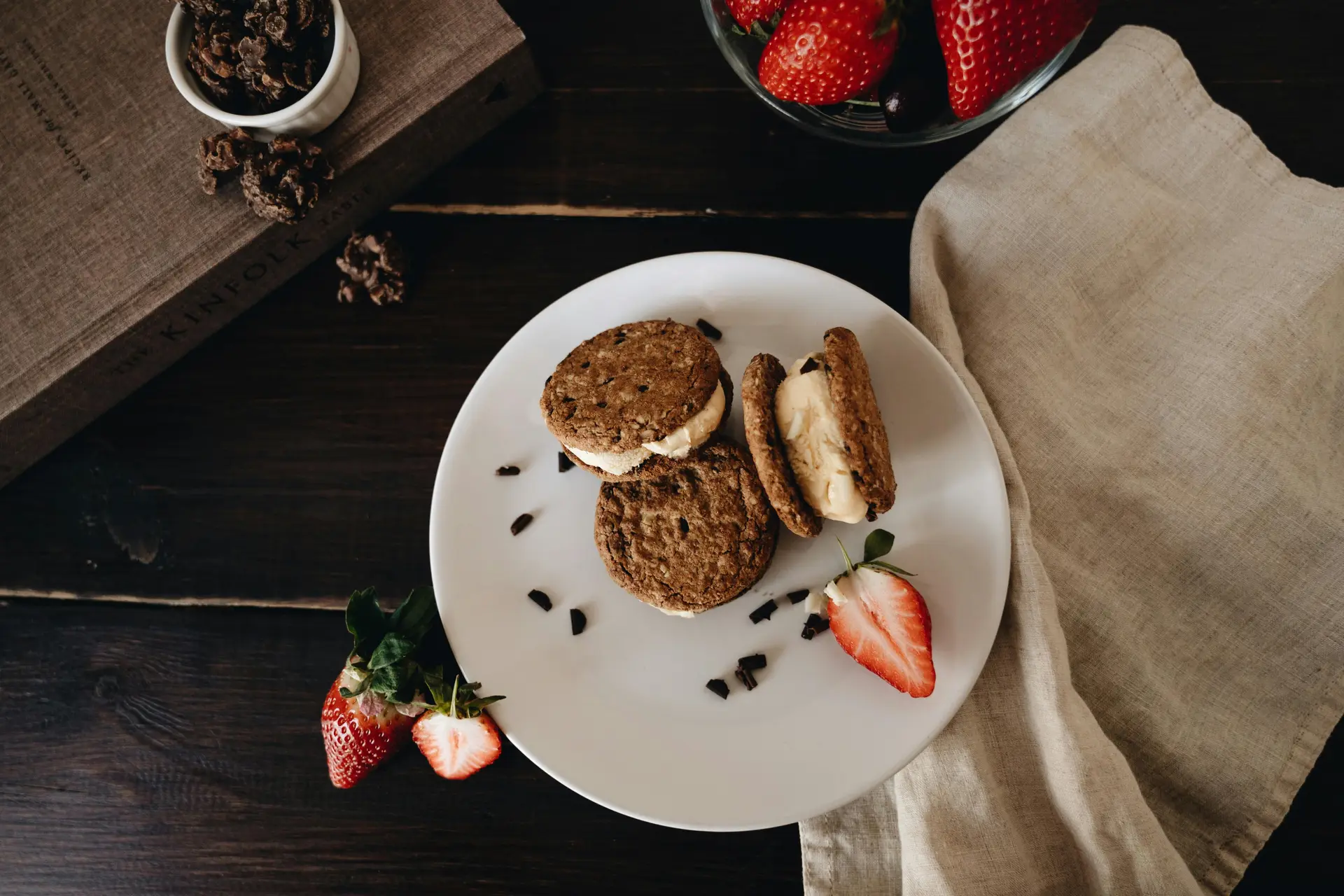 There are some ice cream sandwiches on a white plate along with some strawberries.